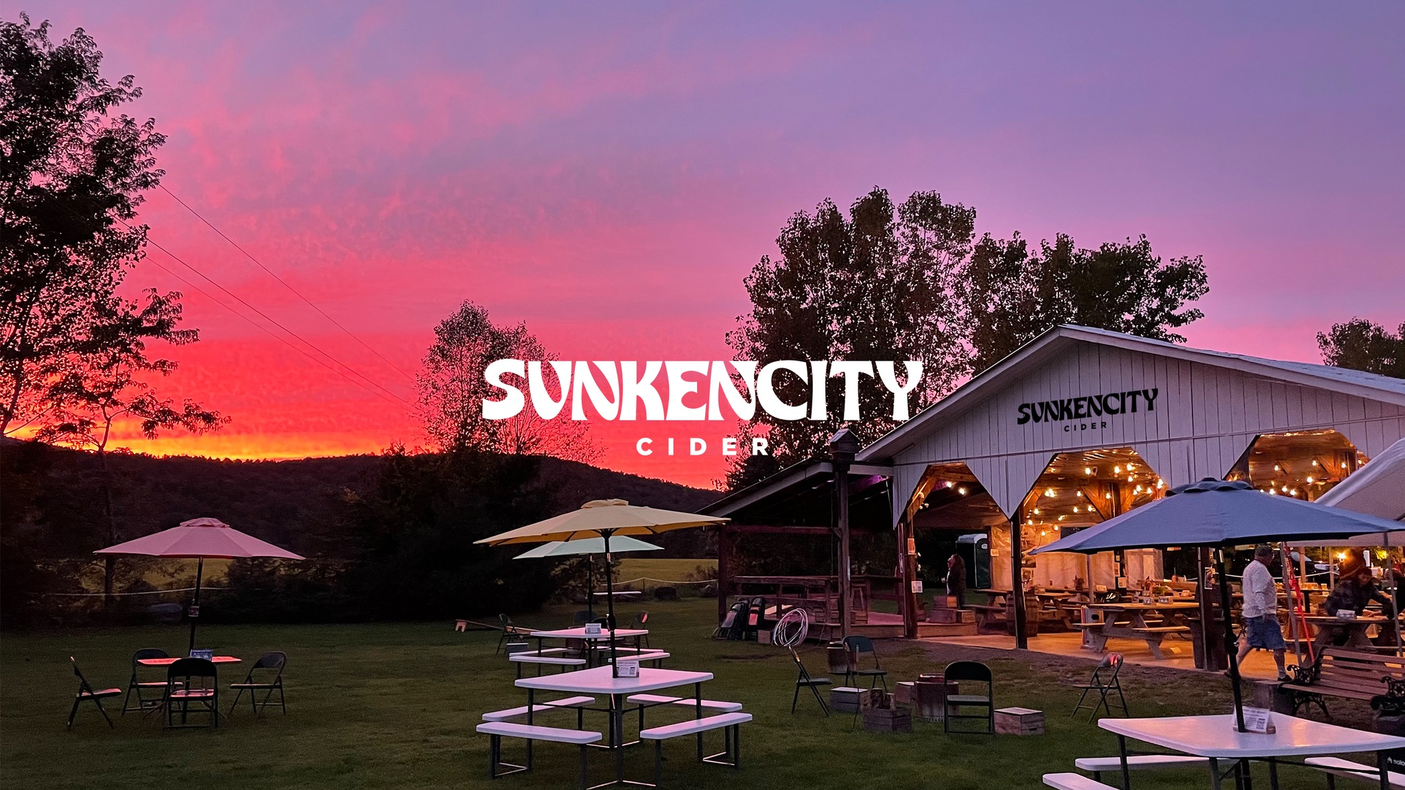 An image of an open barn with the words sunkencity on it in front of a sunset that is pink and purple. there are white picnic tables with umbrellas in the yard in front of it